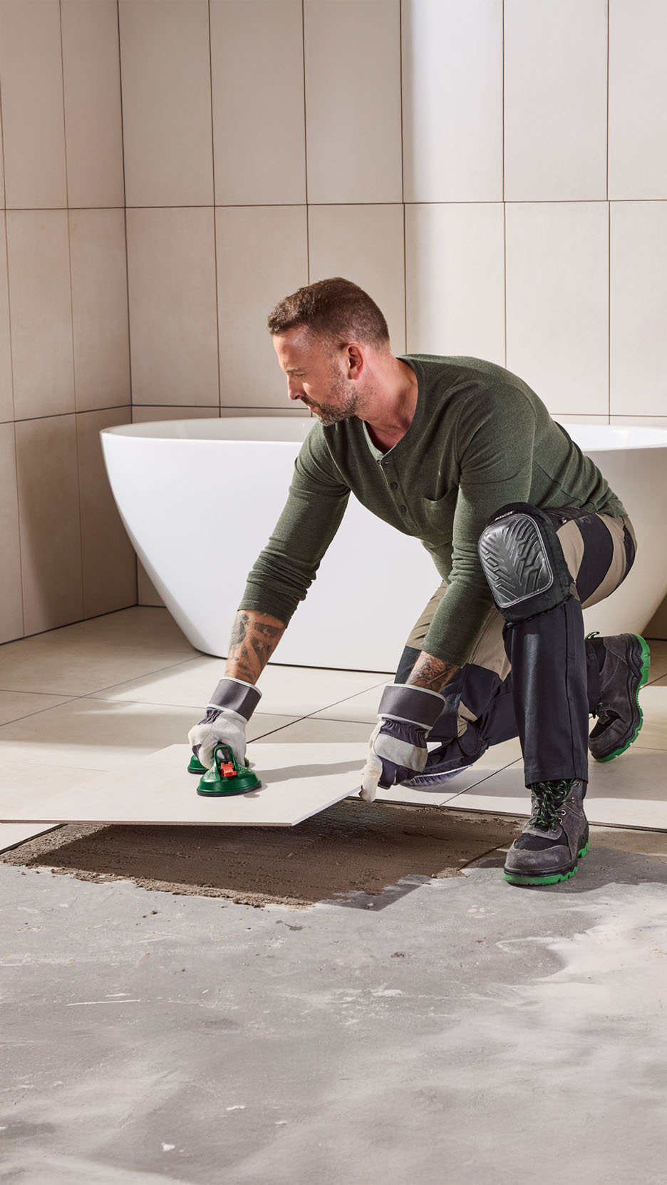 Man laying tiles with a tile suction lifter, wearing work gloves and knee pads.