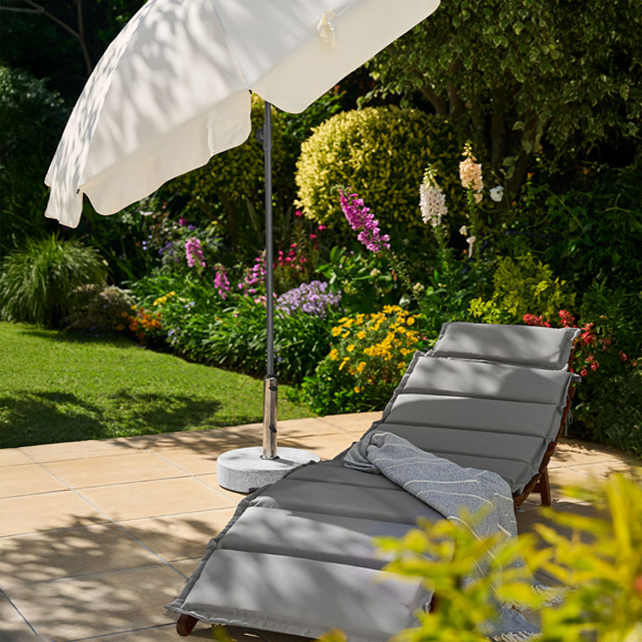 Garden sun lounger with grey cushions and parasol on a patio with a flower bed.