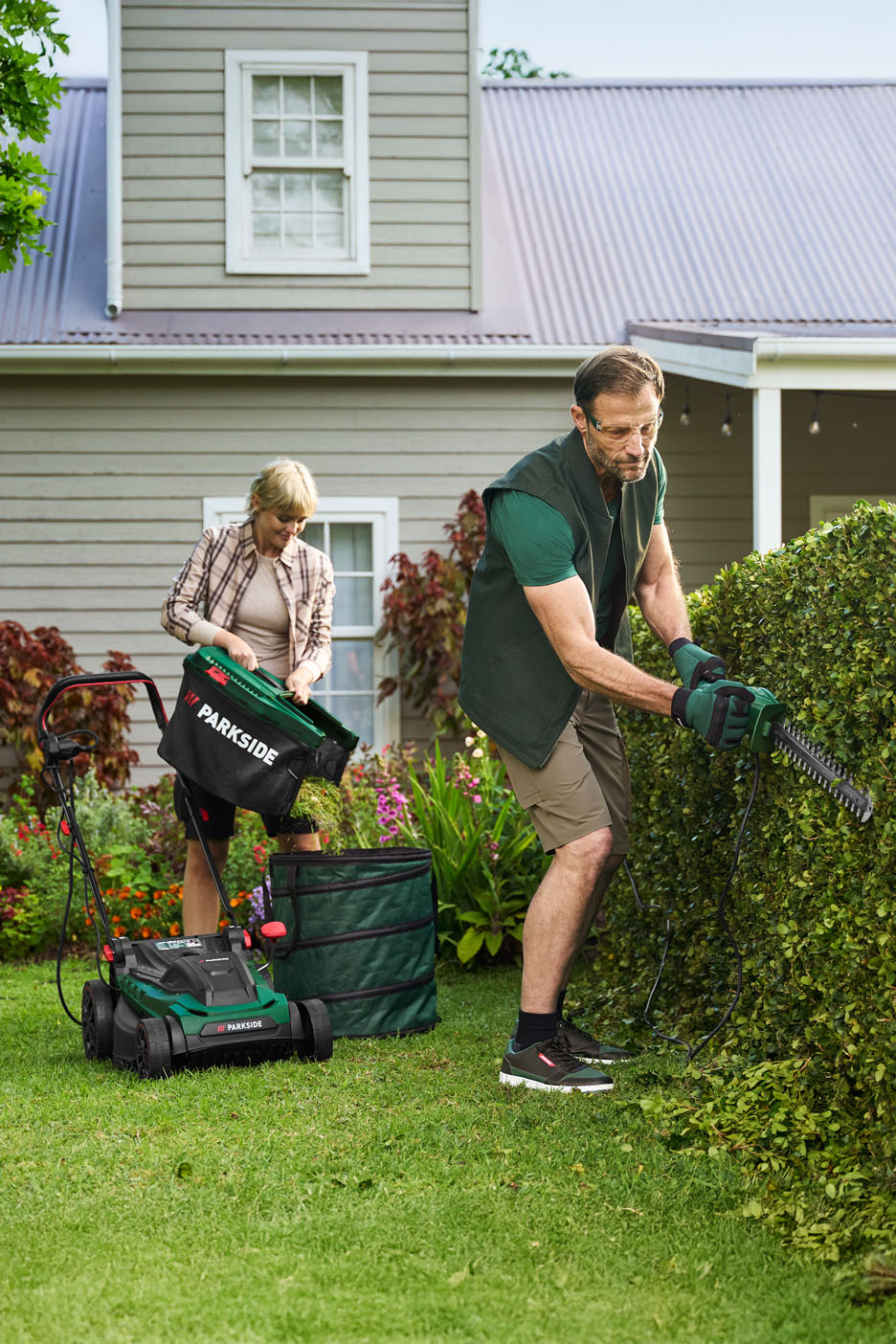 Man trimming hedge, woman emptying lawnmower basket into garden bag.