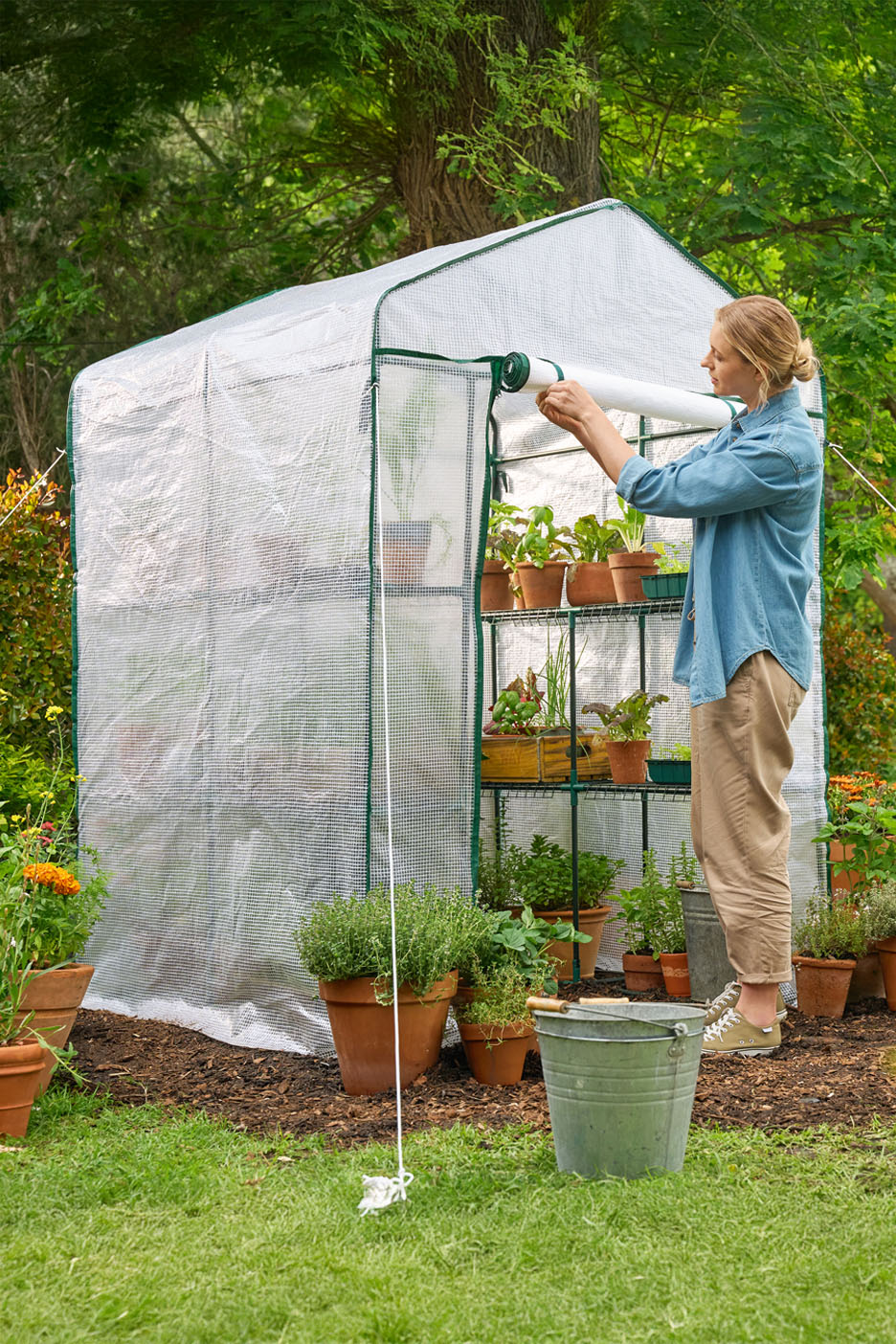 Woman tending to plants in a walk-in greenhouse with shelves of potted herbs and vegetables.