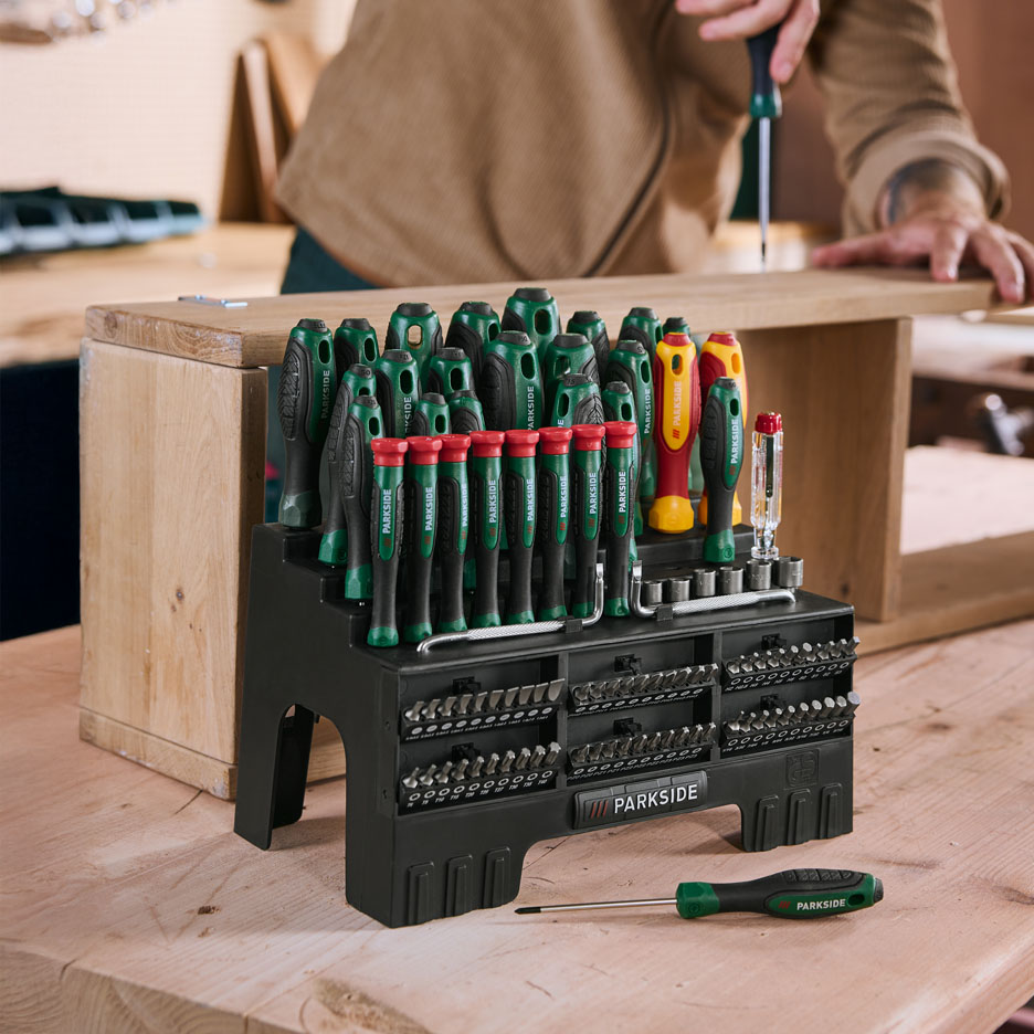 Large set of screwdrivers and bits on a wooden table, with a person working in the background.