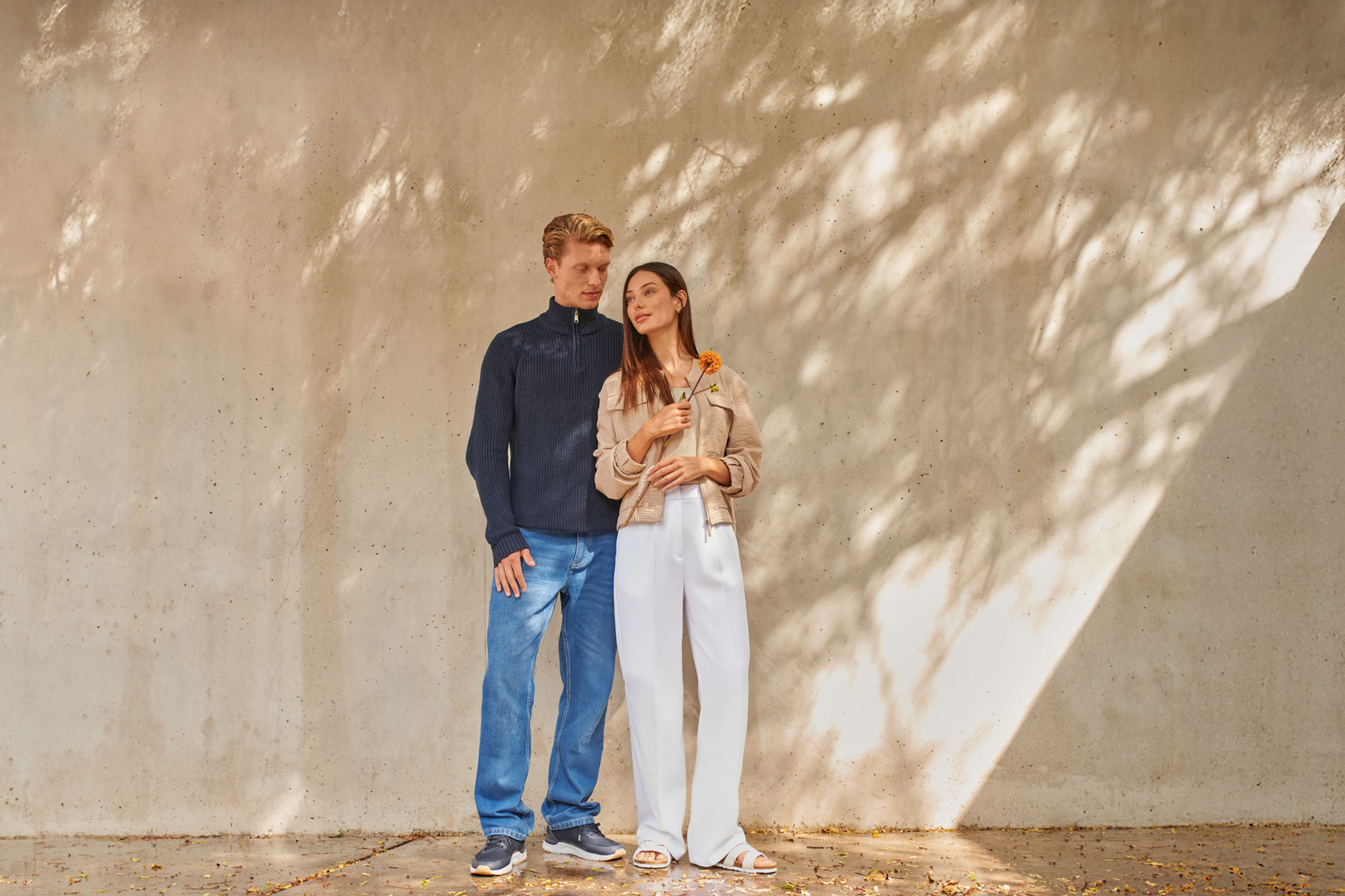 A couple in casual autumn attire: he in a navy sweater and jeans, she in a beige jacket and white pants.