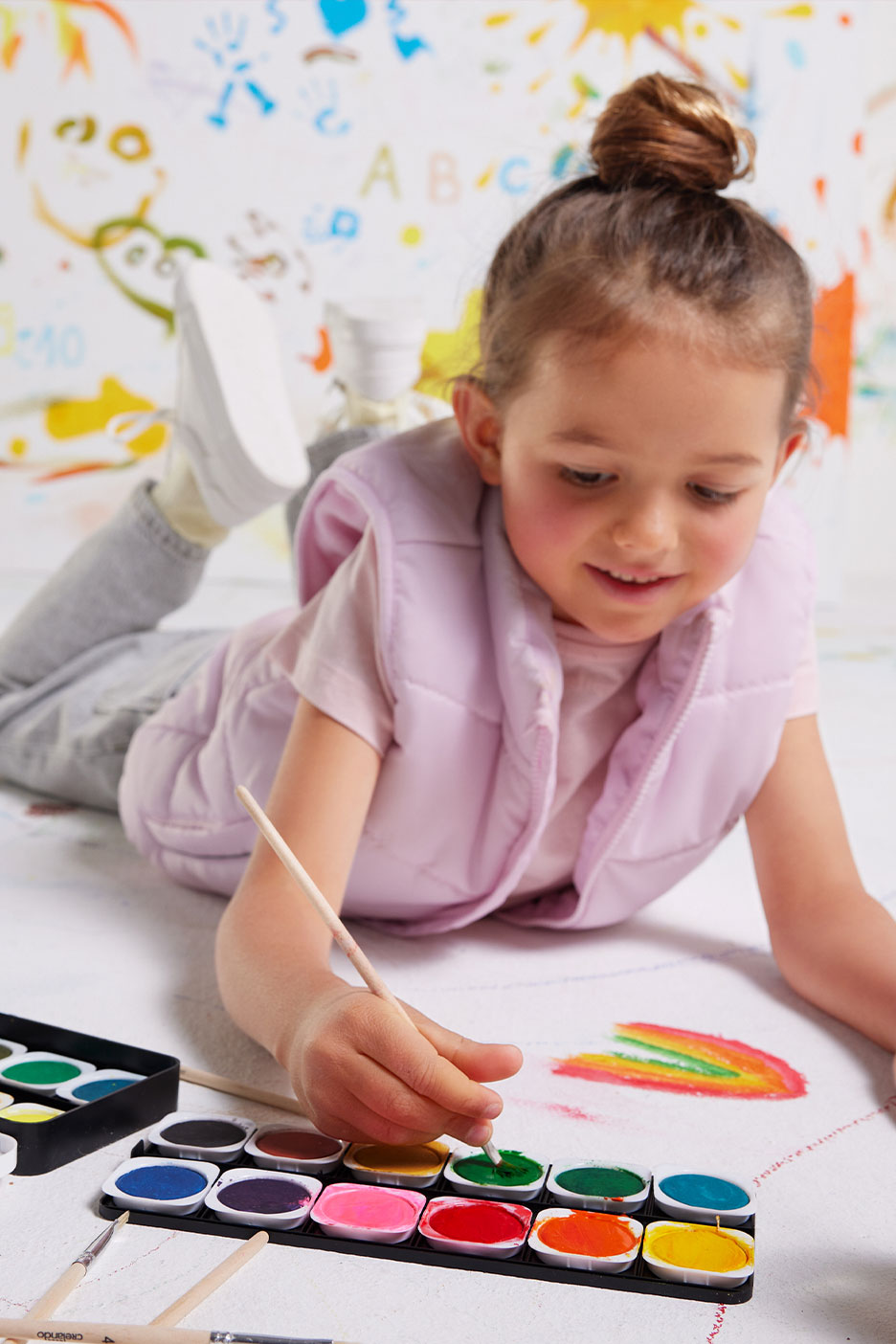 Child painting with watercolors, wearing a pink vest, with colorful drawings in the background.