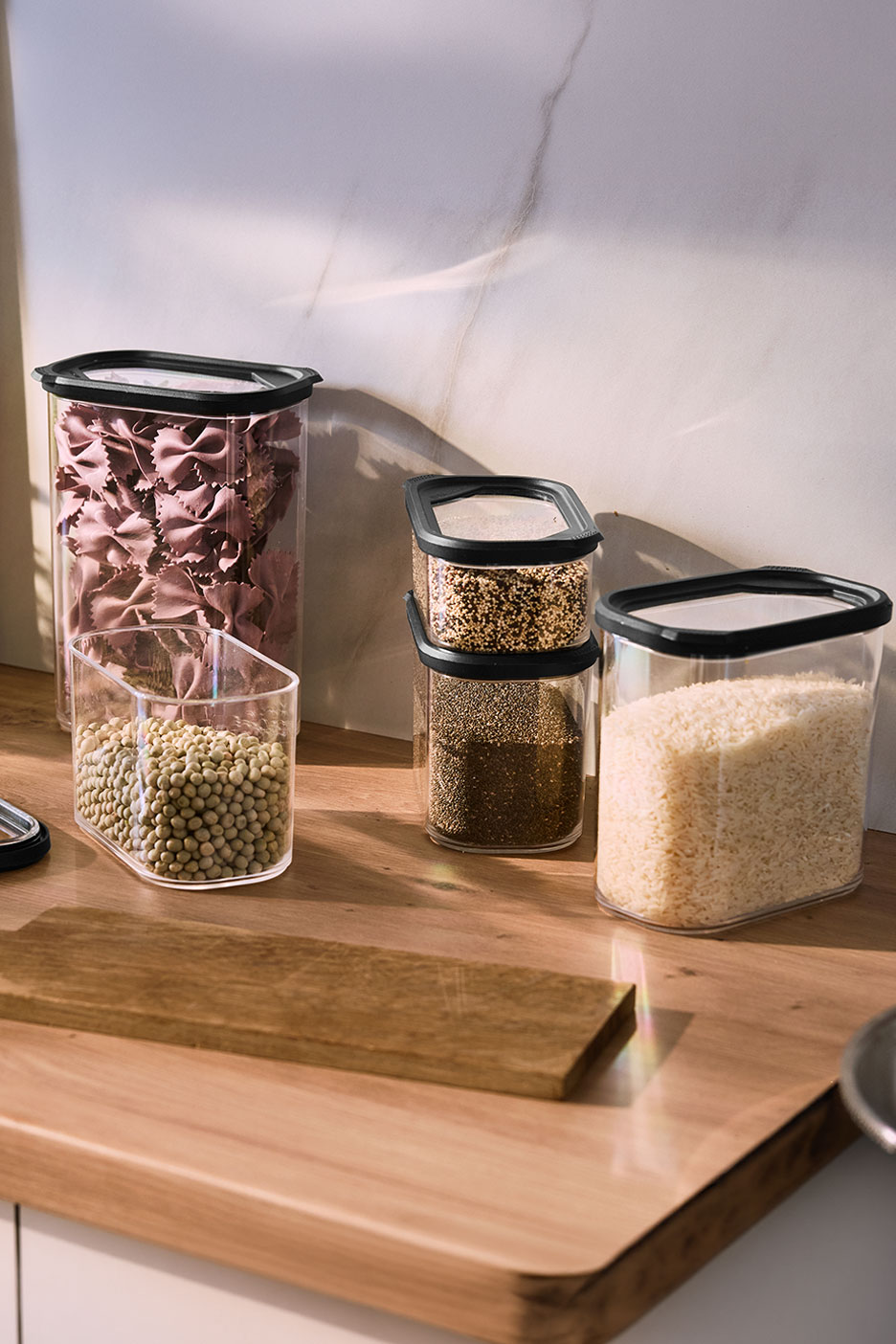 Clear food storage containers with pasta, lentils, quinoa, chia seeds, and rice on a wooden counter.