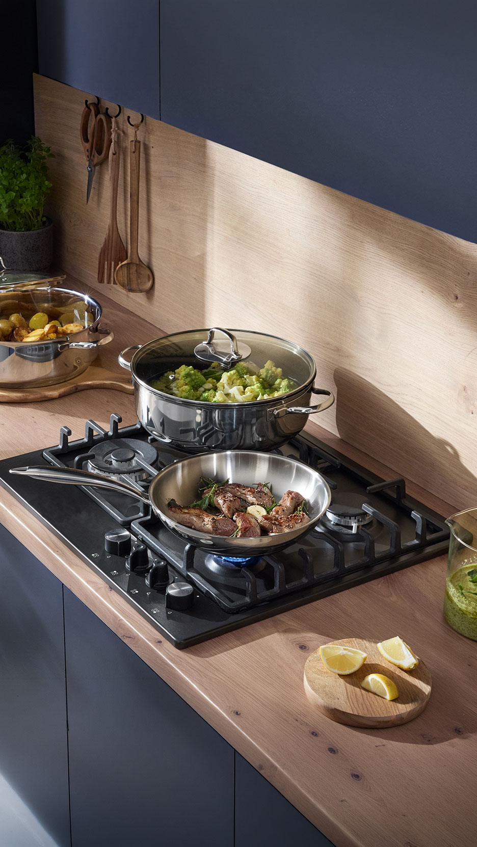 Modern kitchen with gas hob, pans with meat and vegetables, and wooden utensils.