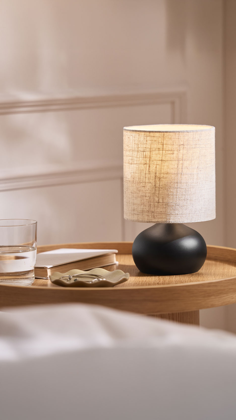 Bedside table with a lit lamp, glass of water, book, and jewelry dish.