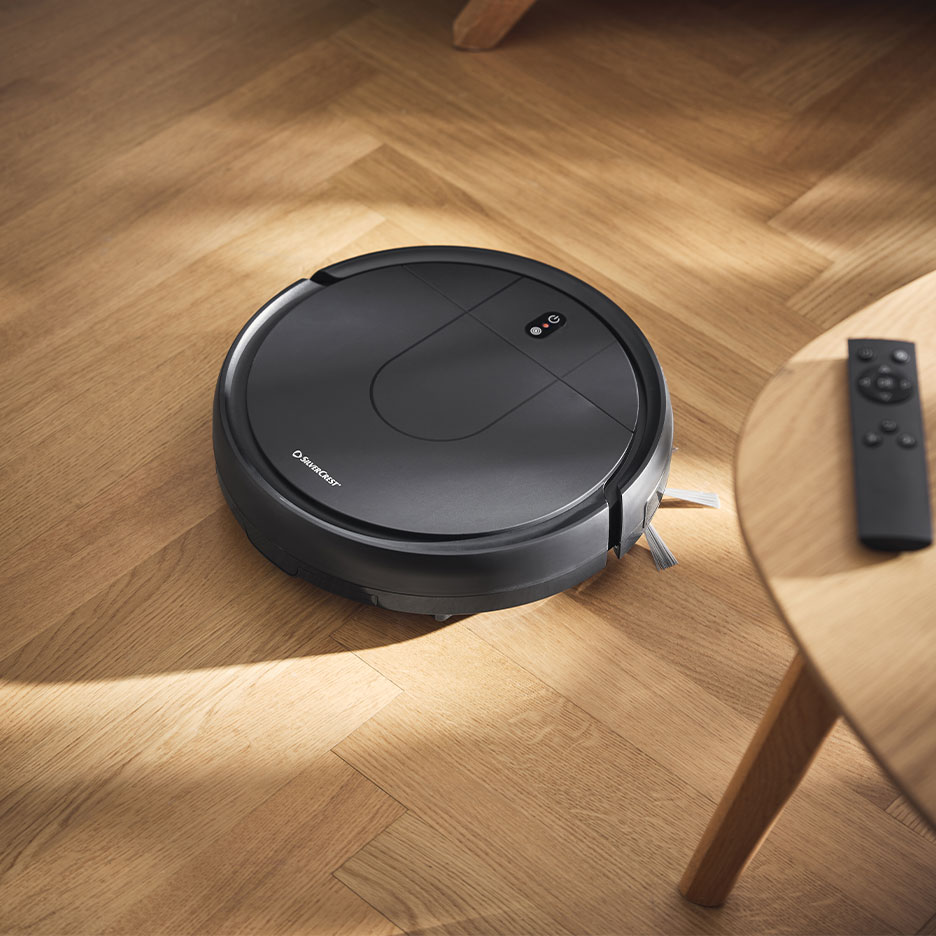 Black robotic vacuum cleaner on a wooden floor next to a small table with a remote control.