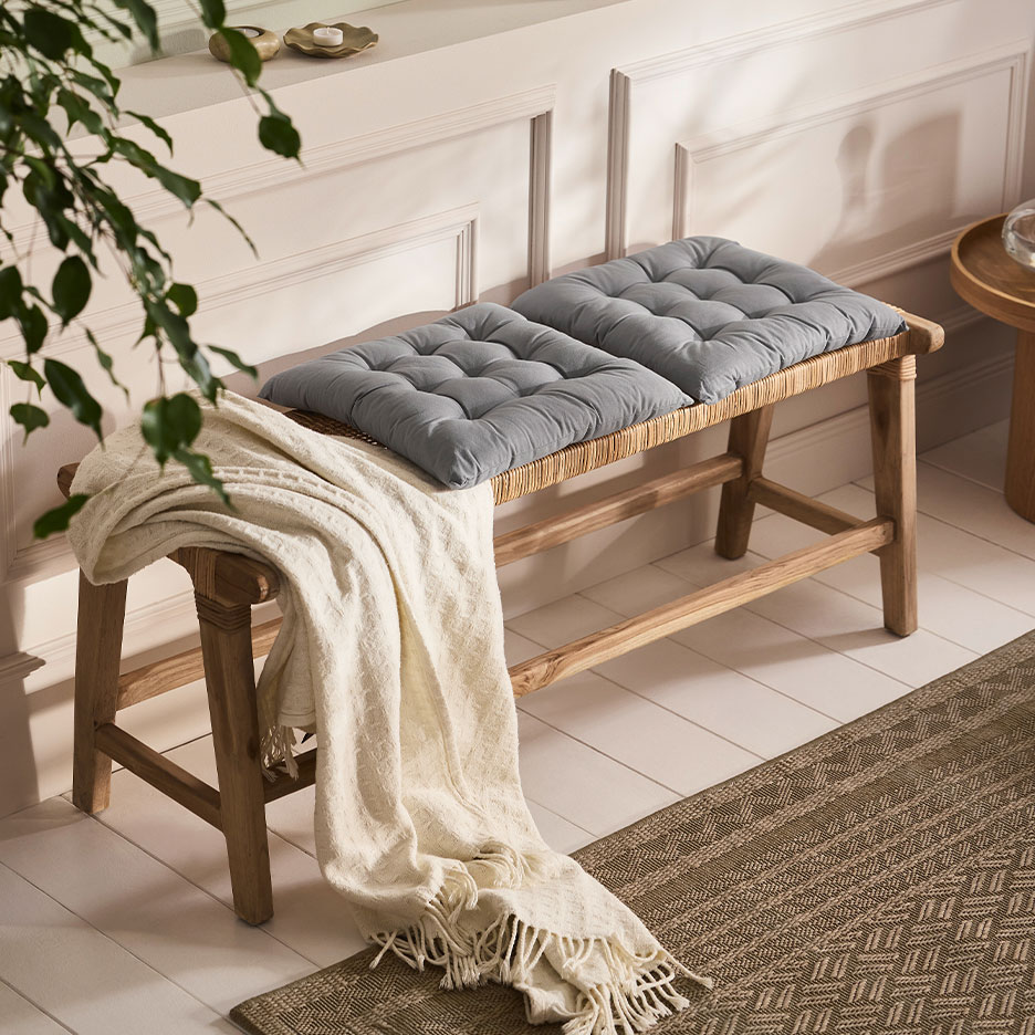 Wooden bench with woven details, grey cushions, and a white throw in a living room setting.
