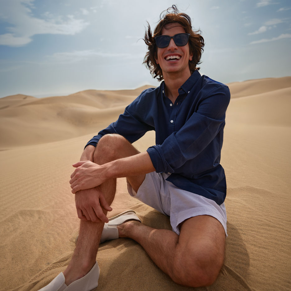 Man in a navy shirt and shorts sits on the dunes, smiling.