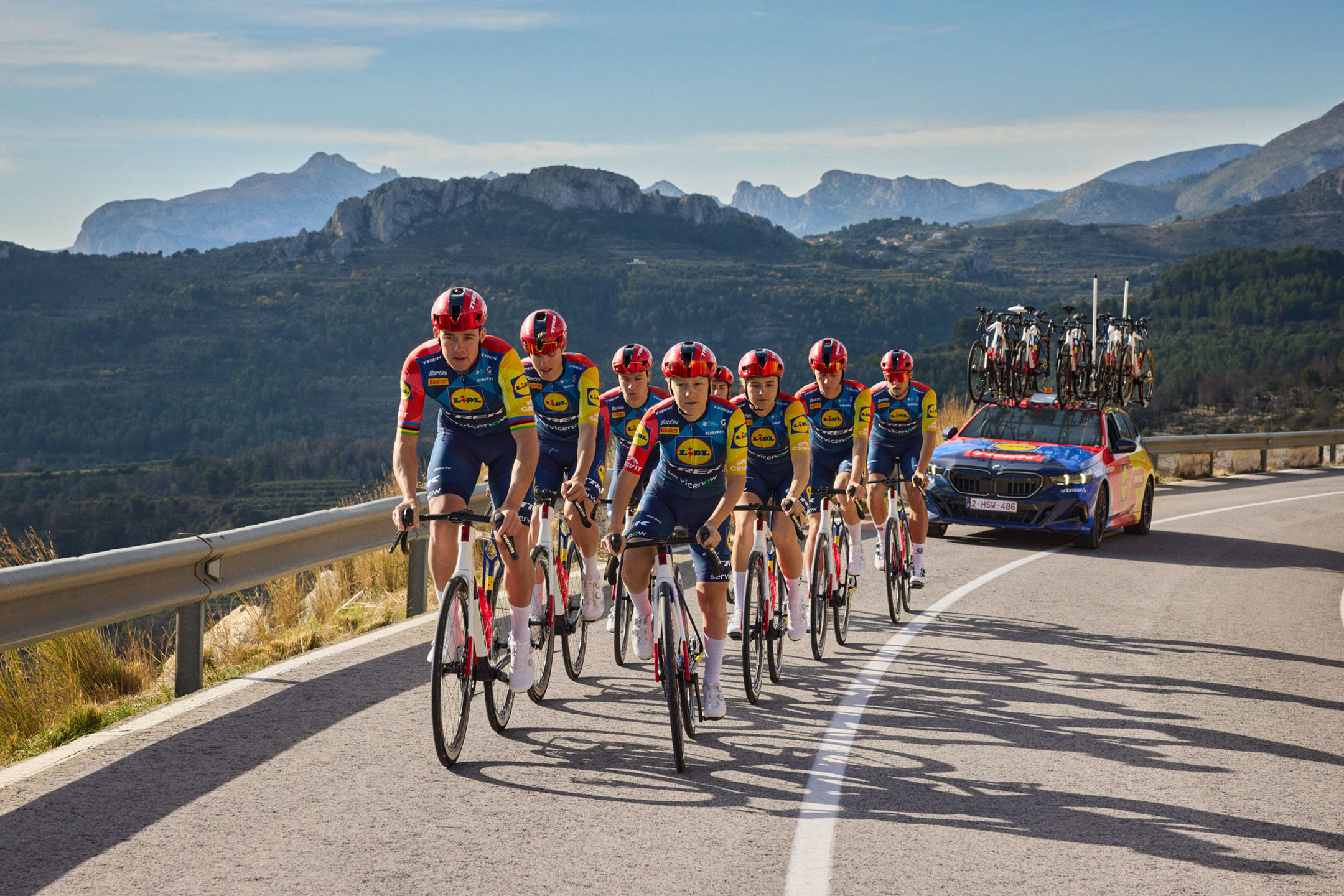 Cyclists in colorful jerseys on a mountain road, with a support car and bicycles on the roof.