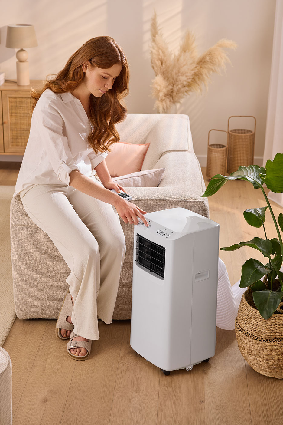 Woman sitting on a sofa, adjusting a white portable air conditioner.