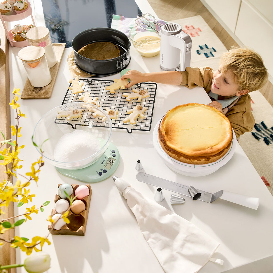 Child decorating cookies and cheesecake with baking tools, scale, and ingredients on a kitchen counter.