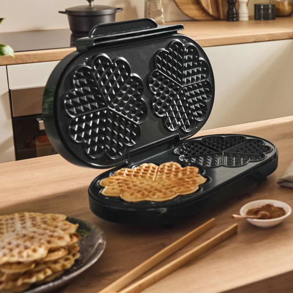 Waffle maker with heart-shaped waffles on a wooden kitchen counter.