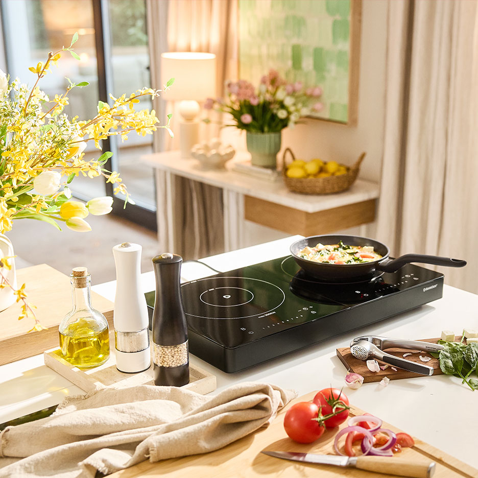 Black double induction hob with a pan, vegetables, and spices on a table.