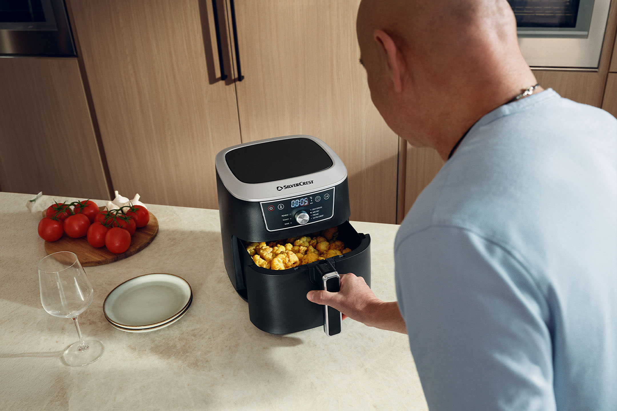 Man checking an air fryer filled with food, with tomatoes and garlic on the counter.