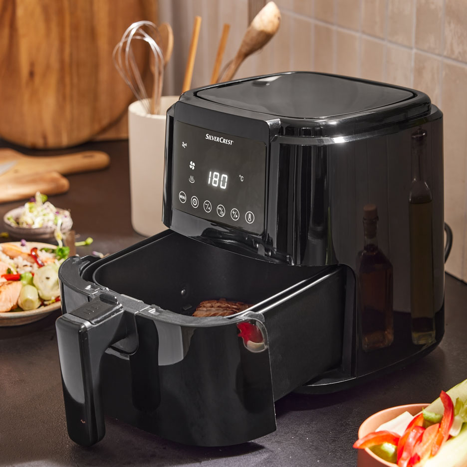 Black air fryer with open basket and meat inside, in a kitchen with food